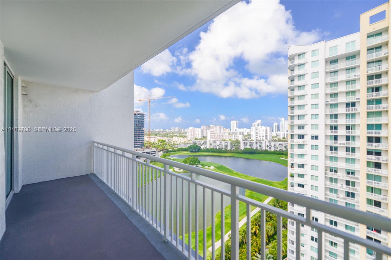 Covered balcony featuring a neutral-toned floor surface and white metal railings overlooking a lake, green space, and urban skyline Covered balcony featuring a neutral-toned floor surface and white metal railings overlooking a lake, green space, and urban skyline