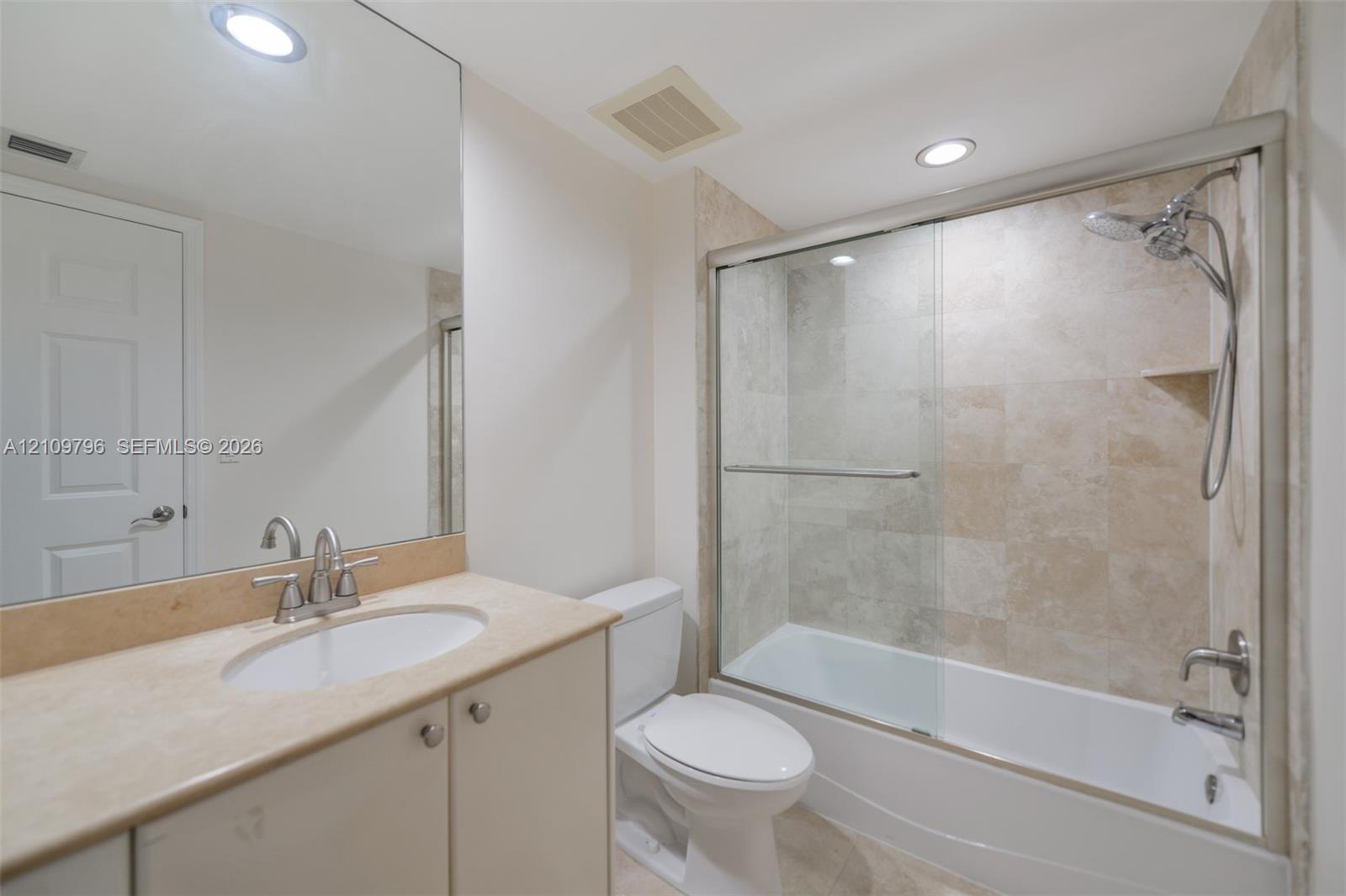 Bathroom featuring a vanity with a stone-finish countertop, undermount sink, full-width mirror, and a shower-tub combination with glass sliding doors and stone-tile wall surround Bathroom featuring a vanity with a stone-finish countertop, undermount sink, full-width mirror, and a shower-tub combination with glass sliding doors and stone-tile wall surround