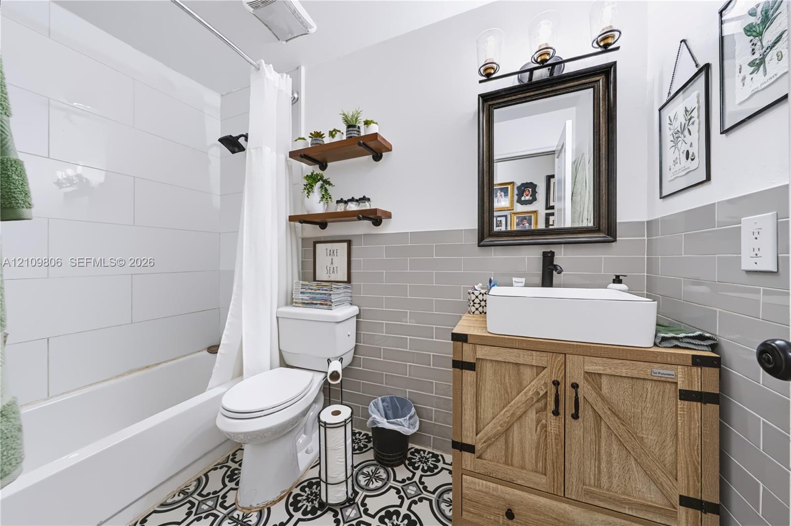 Bathroom featuring wood-finish cabinetry, rectangular vessel sink, matte black hardware, grey subway tile wainscoting, and patterned floor tile