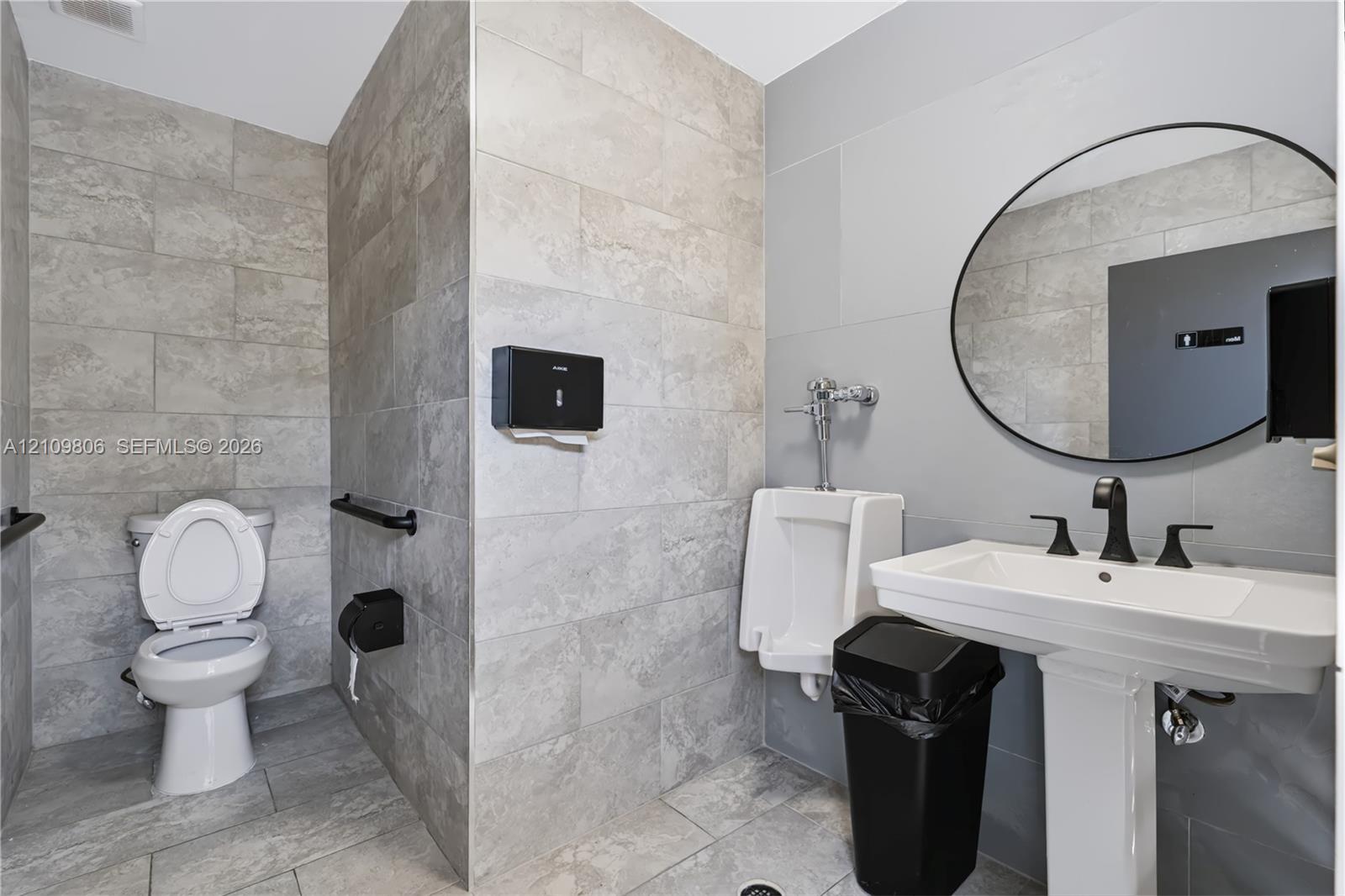 Restroom featuring large-format neutral-toned wall and floor tile, wall-mounted porcelain urinal with manual flushometer, pedestal sink with matte black widespread faucet, and circular frameless mirror