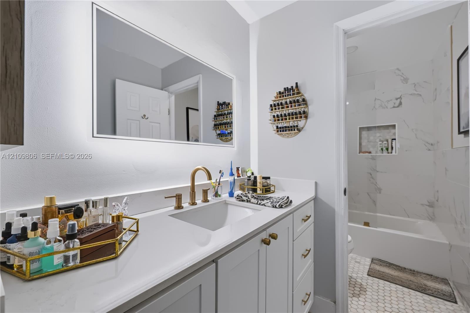 Bathroom featuring a vanity with quartz countertop, undermount sink, and brass-tone faucet hardware, complemented by an integrated backlit mirror and a tiled shower surround with a built-in recessed niche