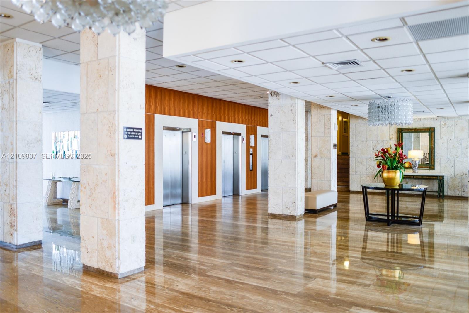 Lobby featuring polished stone flooring, stone-clad structural columns, wood-paneled elevator surround, suspended crystal-style light fixtures, and a recessed drop ceiling