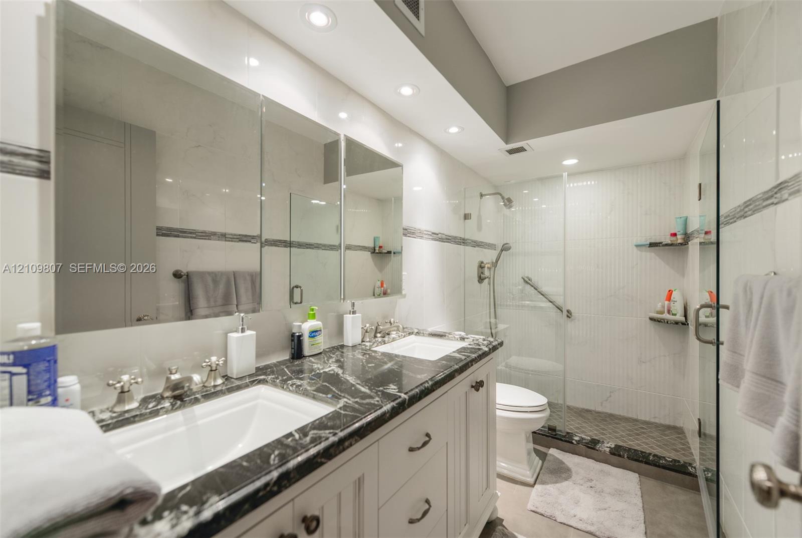 Dual-sink vanity with stone countertop, recessed lighting, expansive wall-to-wall mirror, and glass-enclosed shower with tiled surround