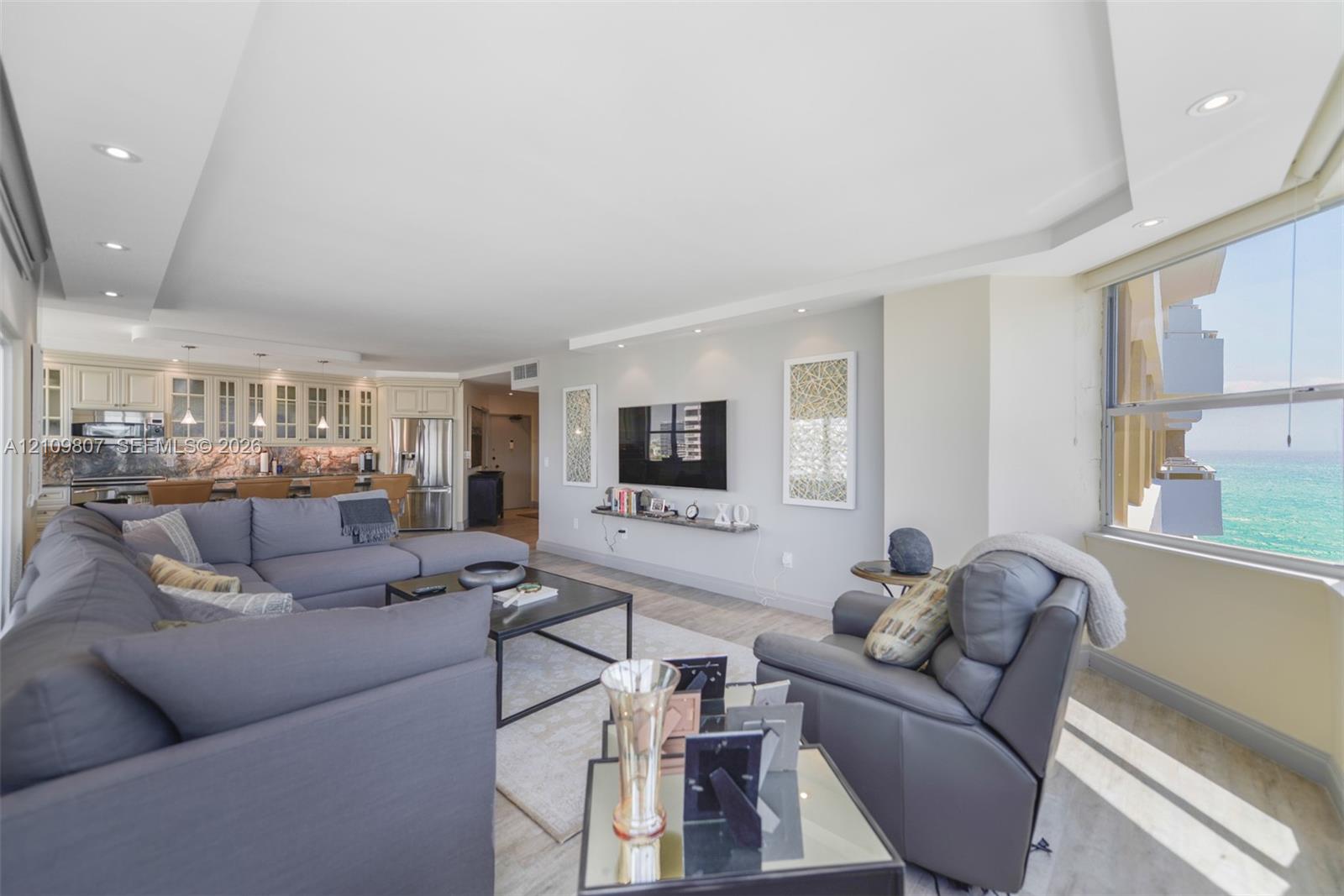 Open-concept living space featuring a tray ceiling with recessed lighting, wood-finish flooring, built-in kitchen cabinetry with glass-front uppers, and oversized windows