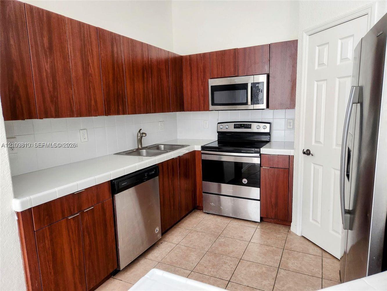 Kitchen featuring wood-finish cabinetry, stainless steel appliances, white tile backsplash, solid-surface countertops, and tile flooring