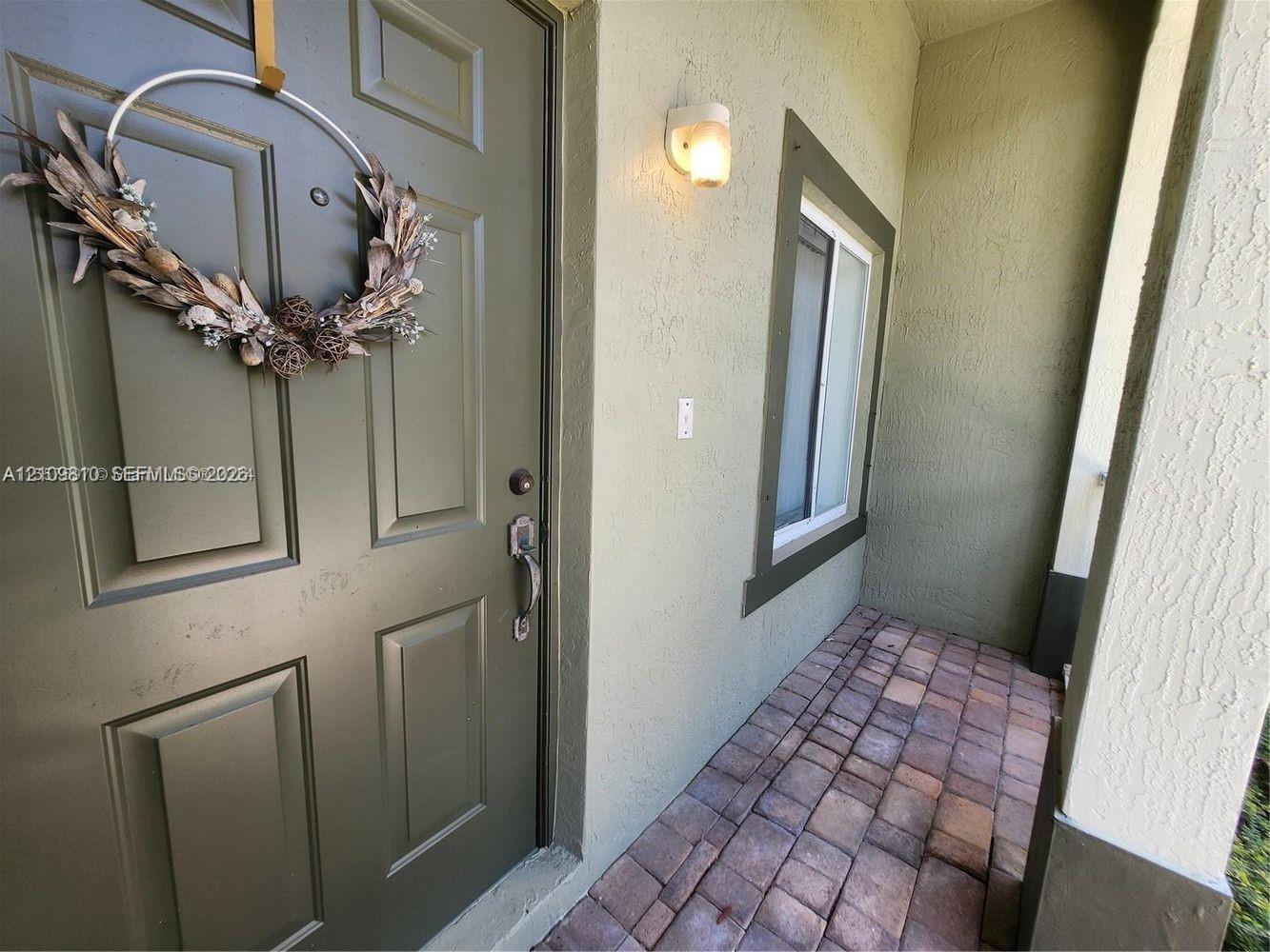 Covered entryway featuring a solid-panel door with metal hardware, paver flooring, and wall-mounted exterior lighting