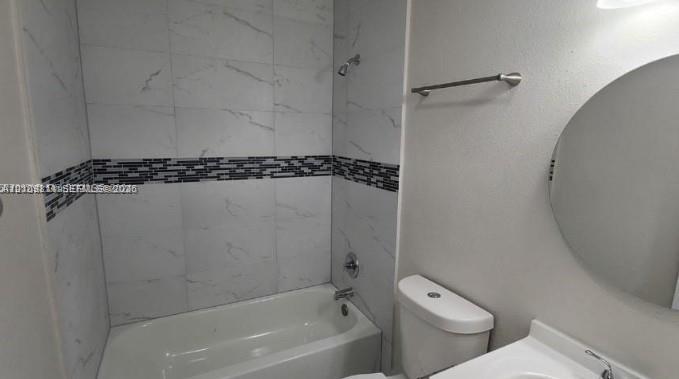 Bathroom featuring a tub-shower combination with large-format stone-pattern tile surround, horizontal mosaic tile accent strip, round wall-mounted mirror, and metal fixtures