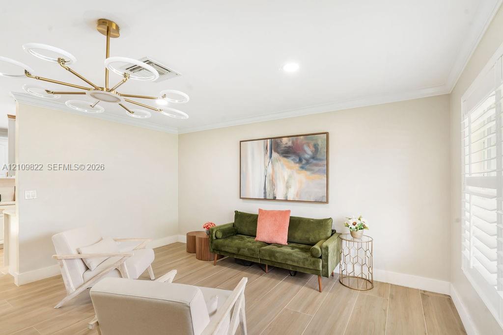Spacious room featuring wood-finish tile flooring, crown molding, tiered gold-tone chandelier with integrated circular lighting, and plantation shutters