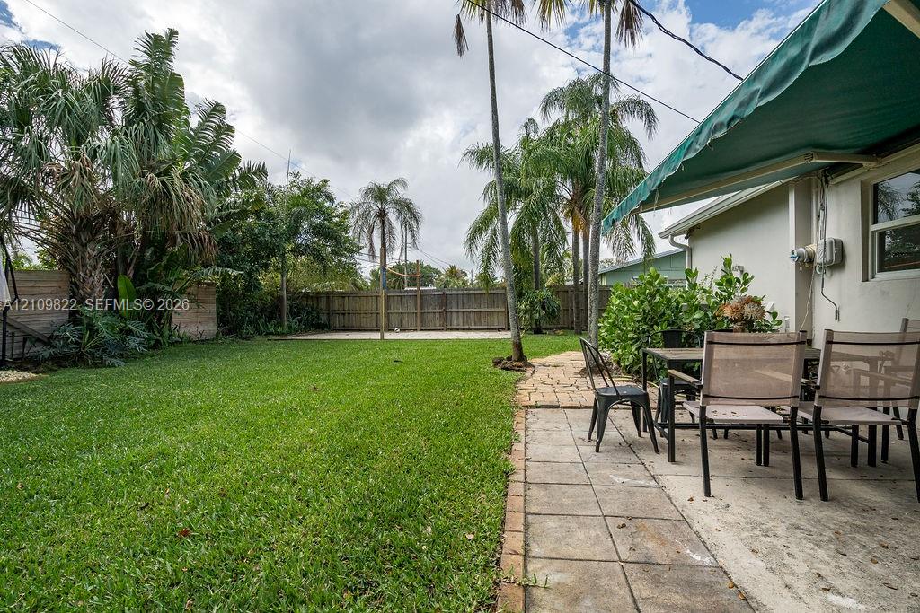 Rear patio featuring a retractable fabric awning, stone paver flooring, perimeter wood fencing, and mature palm trees