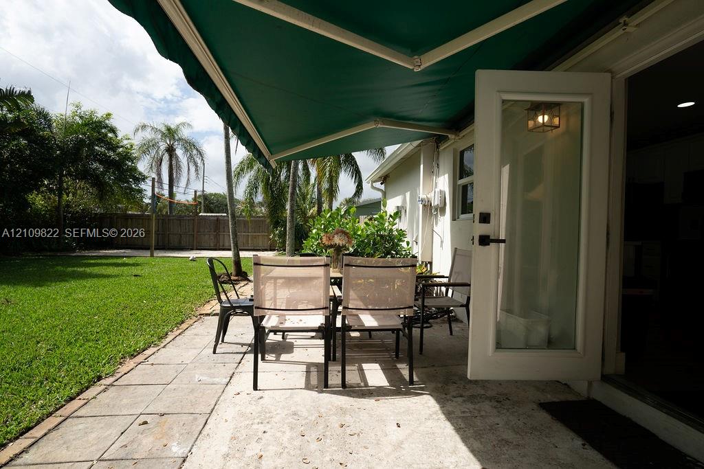 Paved stone patio featuring a retractable green awning, glass-paneled entry door, fenced perimeter, and mature palm landscaping
