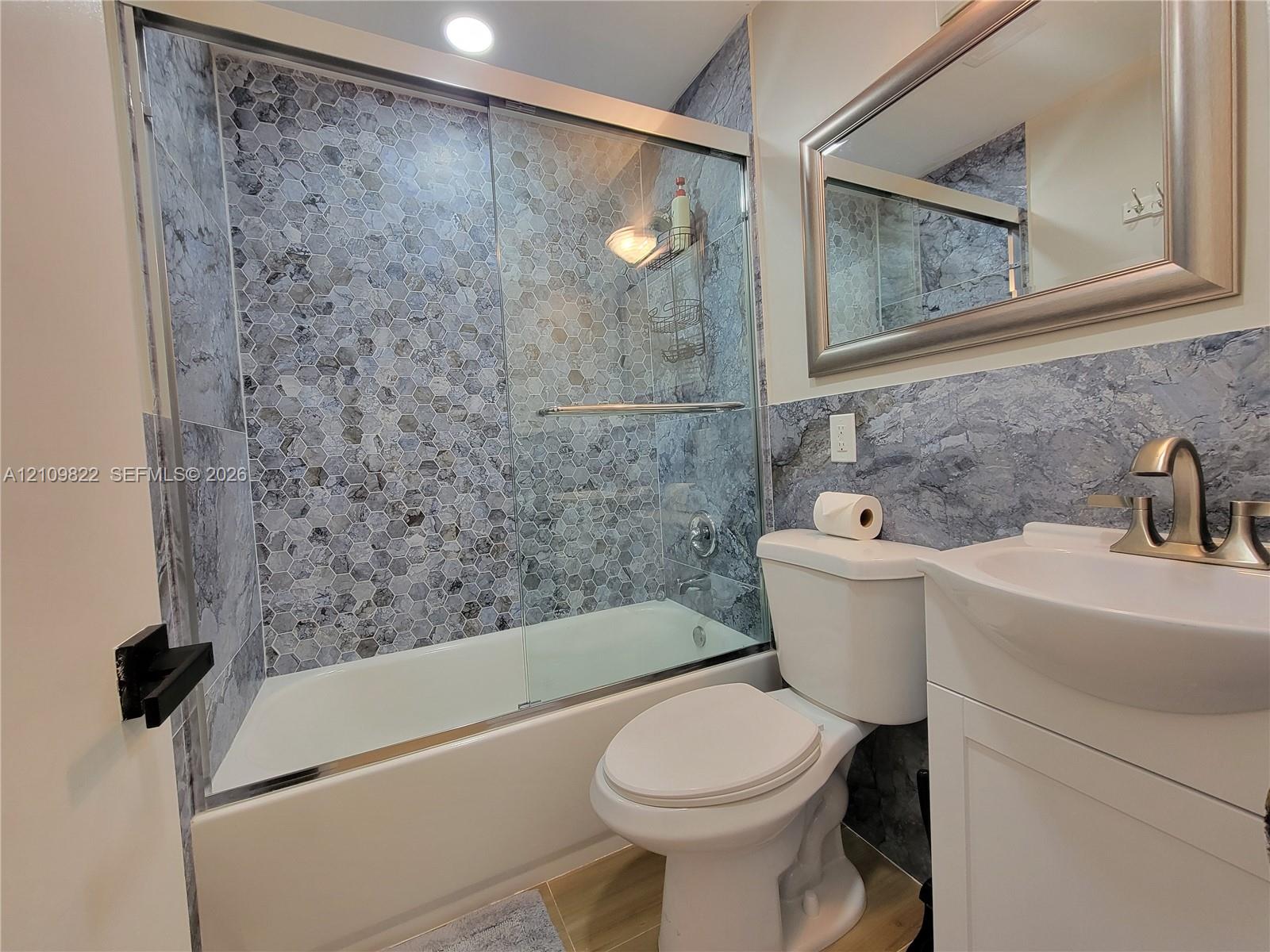 Full bathroom featuring a tub-shower combination with glass sliding doors, honeycomb-patterned tile surround, vanity with an integrated basin, brushed nickel faucet, and a wall-mounted mirror