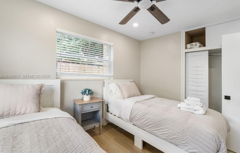 Neutral-toned room featuring wood-finish flooring, a ceiling fan with integrated lighting, a window with horizontal blinds, and a built-in closet with slatted doors and overhead shelving