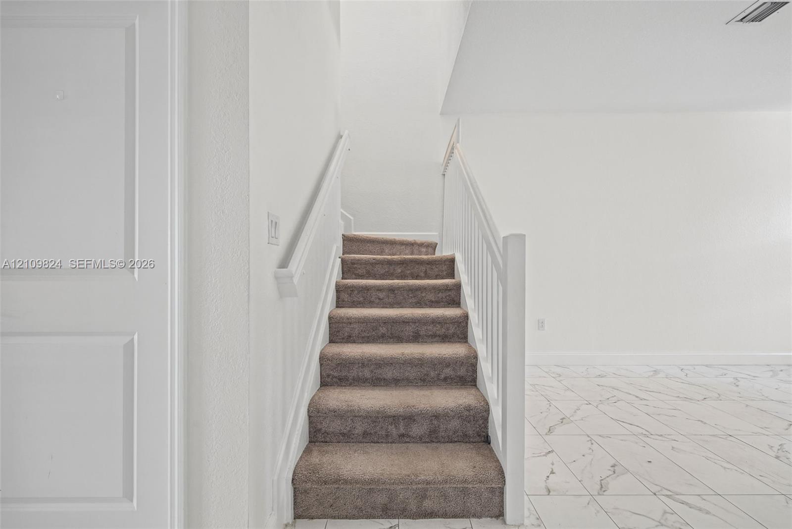 Staircase featuring carpeted steps, white balustrades, and a panel-style door, complemented by large-format tile flooring Staircase featuring carpeted steps, white balustrades, and a panel-style door, complemented by large-format tile flooring