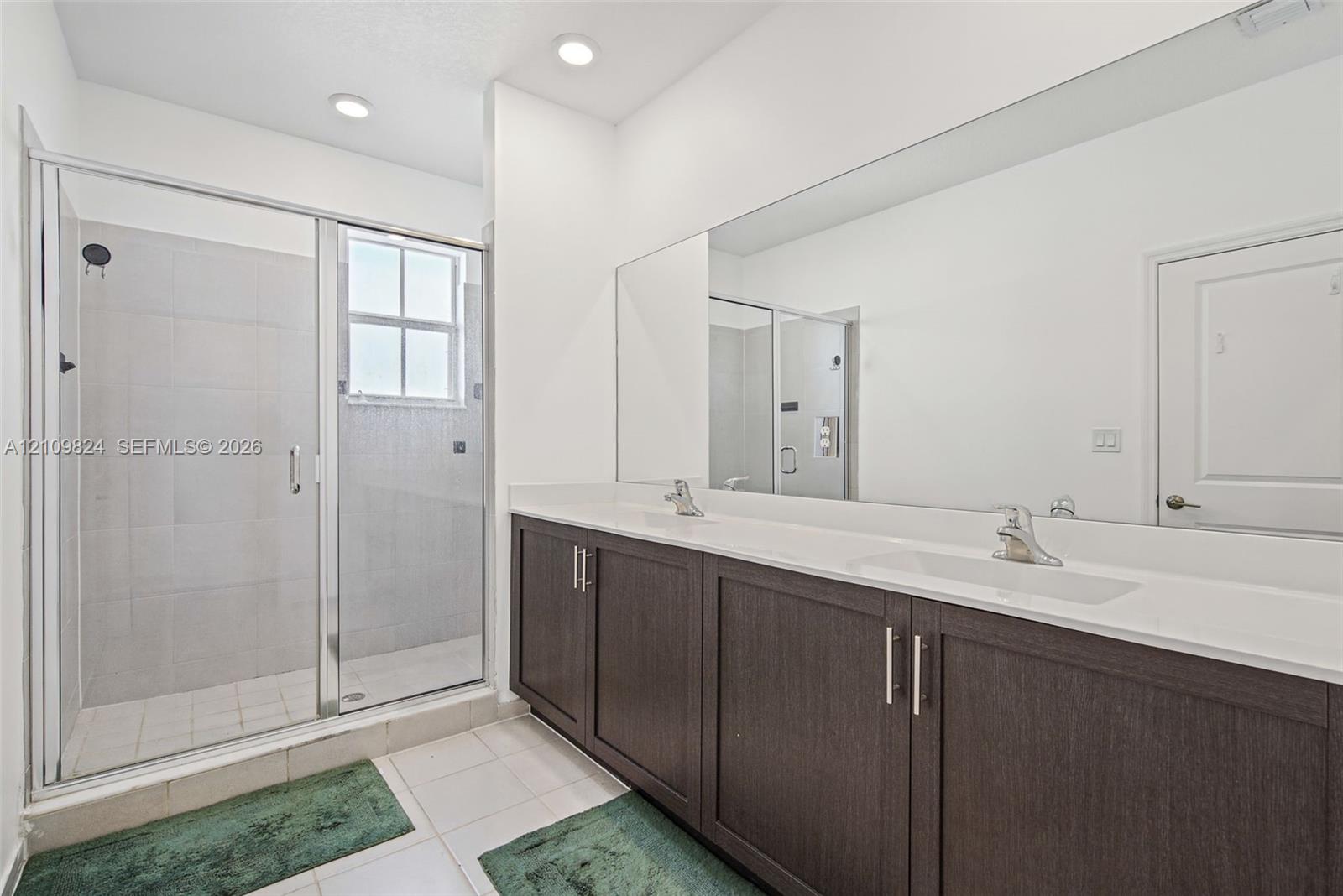 Double vanity with dark-finish cabinetry, solid-surface countertop, full-width wall mirror, and glass-enclosed shower with tiled surround