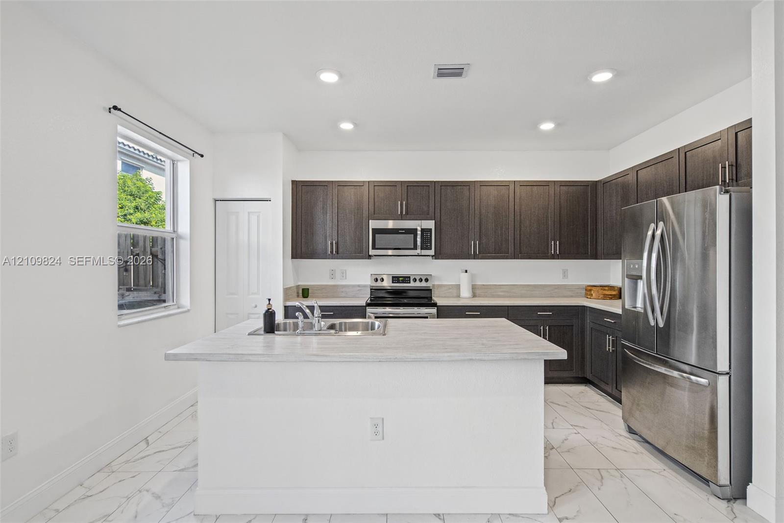 Kitchen featuring dark-toned cabinetry, a central island with a stone-finish countertop, stainless steel appliances, and tile flooring Kitchen featuring dark-toned cabinetry, a central island with a stone-finish countertop, stainless steel appliances, and tile flooring