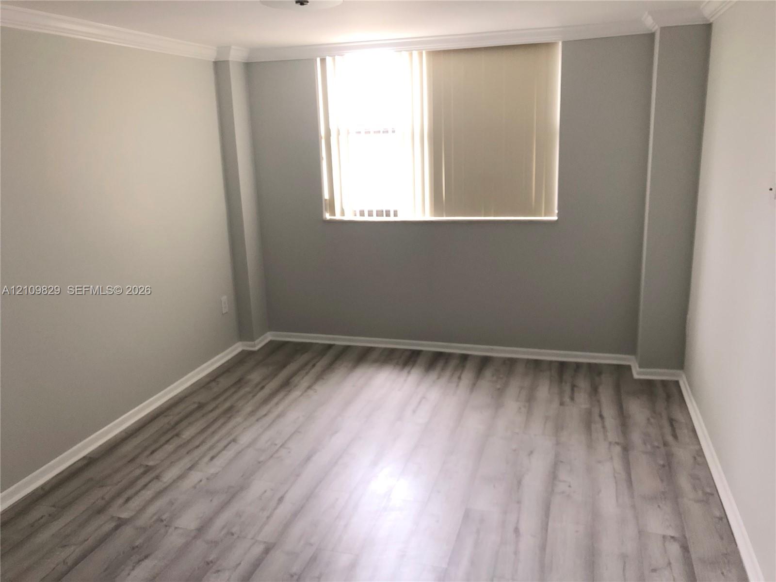 Neutral-toned interior featuring wood-finish flooring, crown molding, and a single window with vertical blinds