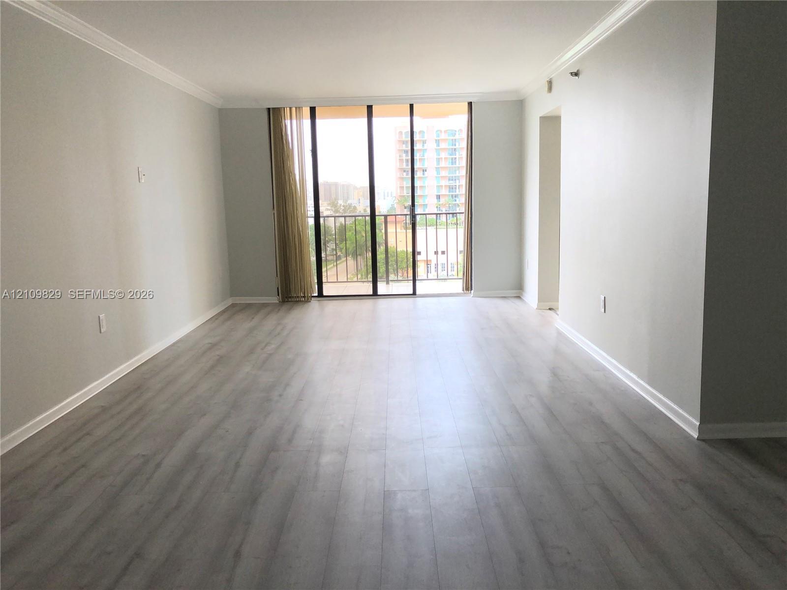 Open-concept room featuring light-toned wood-finish flooring, neutral wall paint, crown molding, and floor-to-ceiling glass sliding doors opening to a balcony