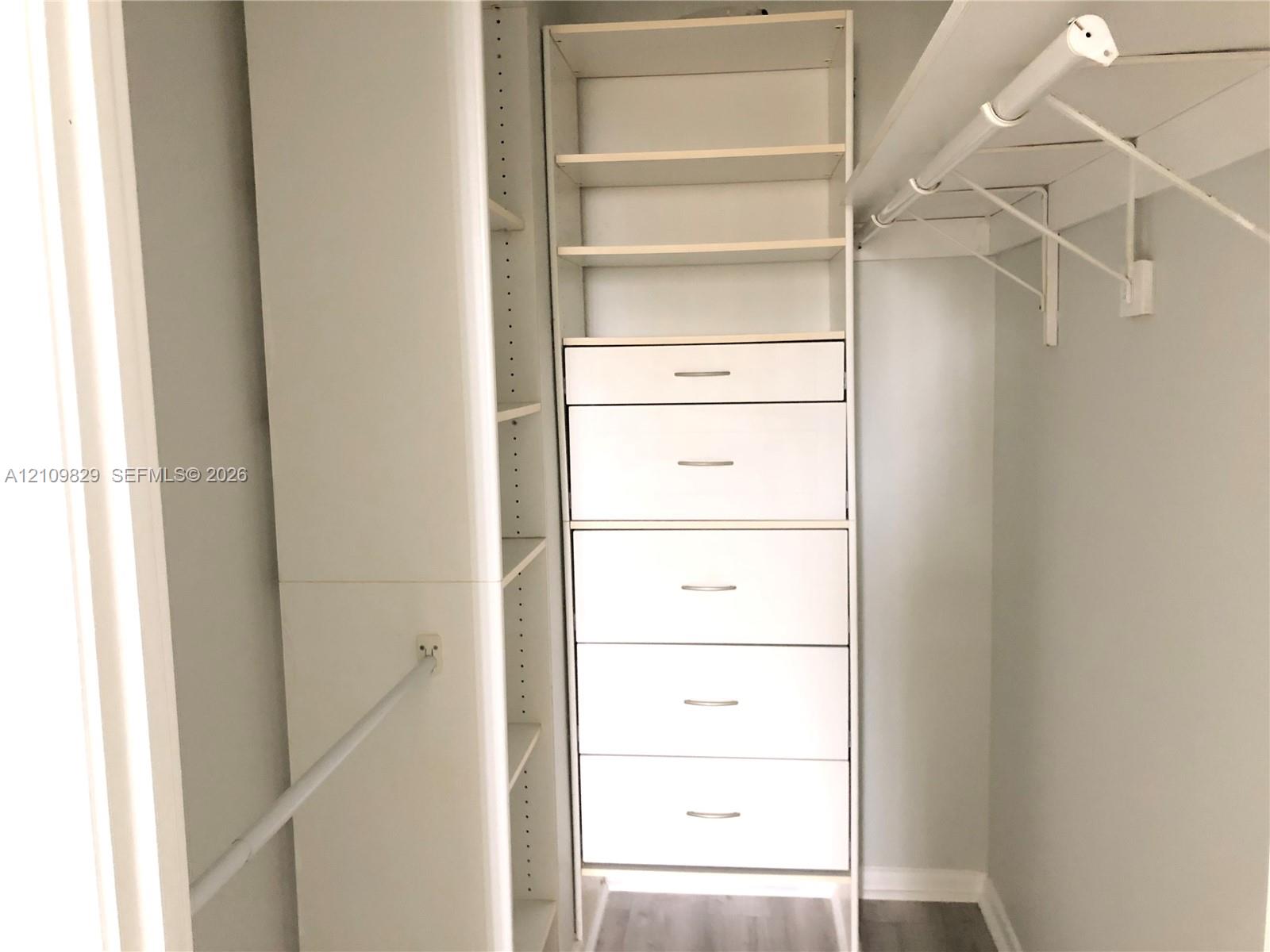 Walk-in closet featuring white built-in cabinetry, five integrated drawers, open shelving, and a wall-mounted garment rod over wood-finish flooring