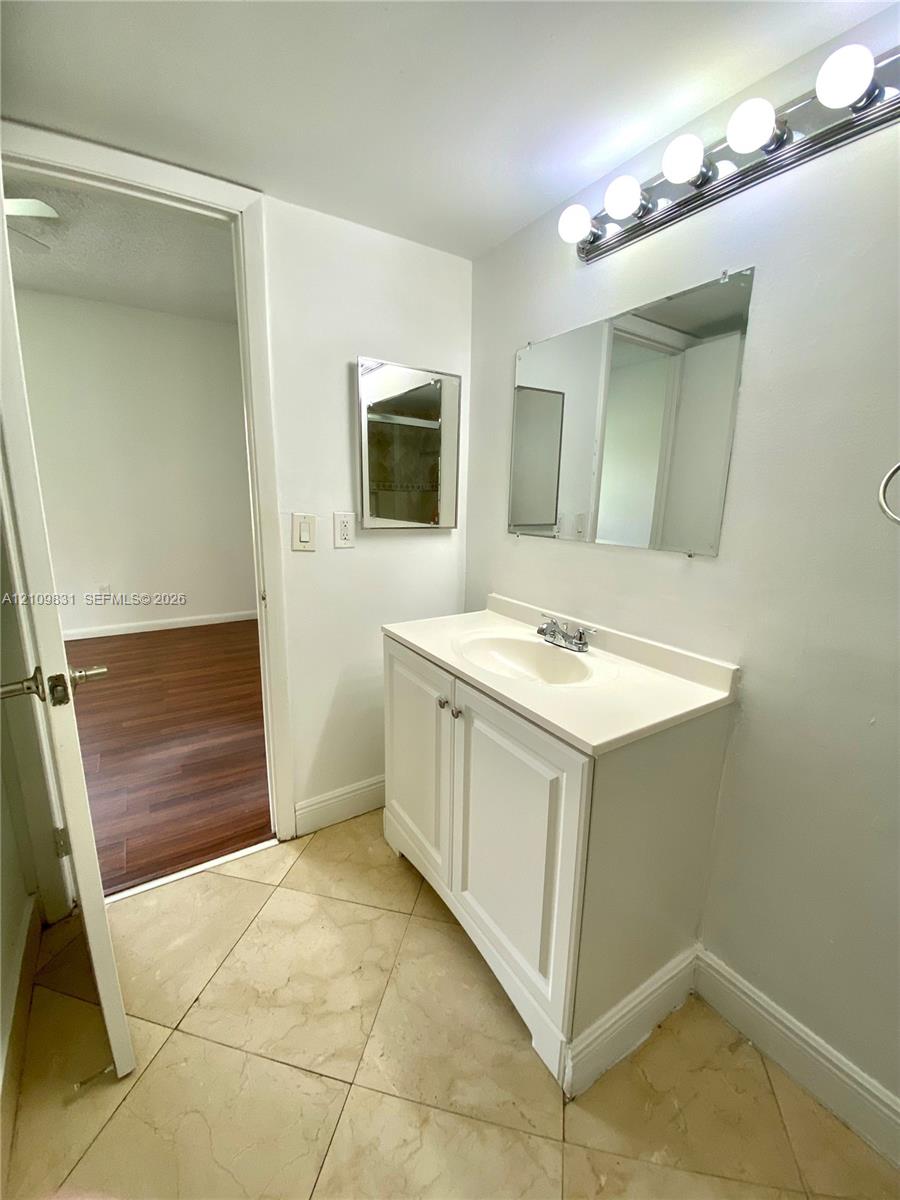 Bathroom vanity featuring a white cabinet base, integrated countertop and sink, wall-mounted multi-bulb lighting fixture, rectangular mirror, and neutral-tone tile flooring
