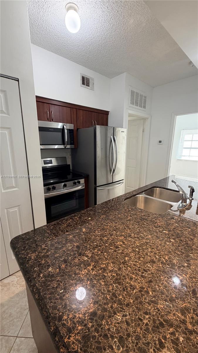 Kitchen featuring dark-toned stone countertops, stainless steel appliances, wood-finish cabinetry, and a dual-basin undermount sink with a gooseneck faucet