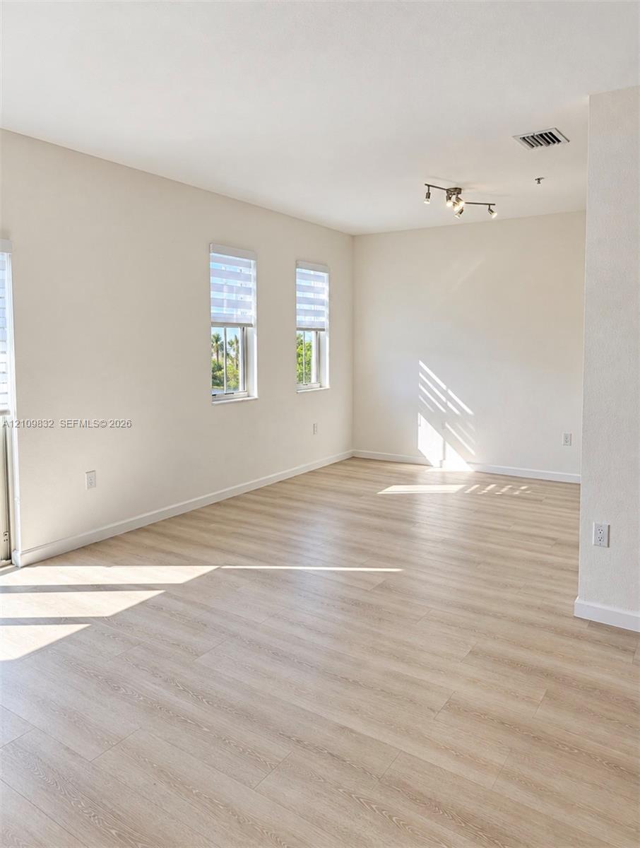 Neutral interior space featuring wood-finish flooring, crisp white walls, dual windows with roller shades, and a multi-directional track lighting fixture