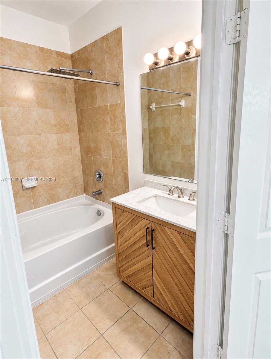Bathroom featuring a wood-finish vanity with a stone countertop, an integrated under-mount sink, a tile-surround soaking tub, and wall-mounted light fixtures