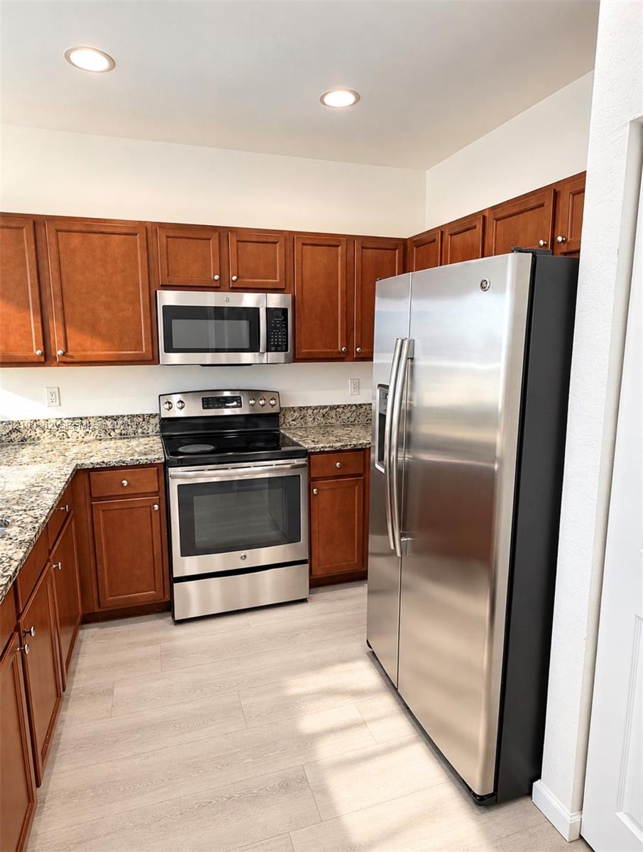 Kitchen featuring stained wood cabinetry, stone-finish countertops, stainless steel appliances, and light-toned wood-finish flooring Kitchen featuring stained wood cabinetry, stone-finish countertops, stainless steel appliances, and light-toned wood-finish flooring
