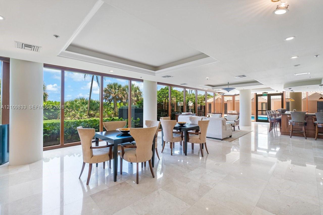 Open-concept interior featuring polished stone flooring, recessed tray ceilings, floor-to-ceiling windows with wood-finish frames, and a built-in wet bar