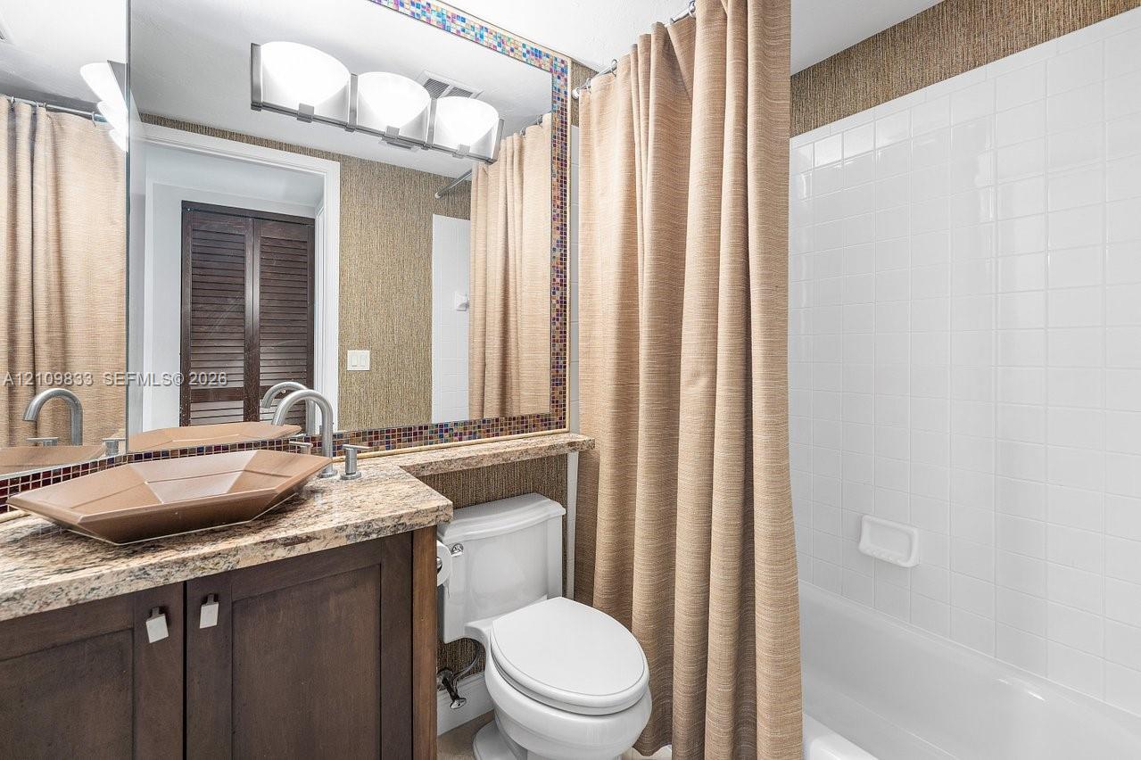 Bathroom vanity featuring stone-finish countertop, geometric vessel sink, dark wood-tone cabinetry, tiled tub surround, and framed mirror with mosaic tile accents