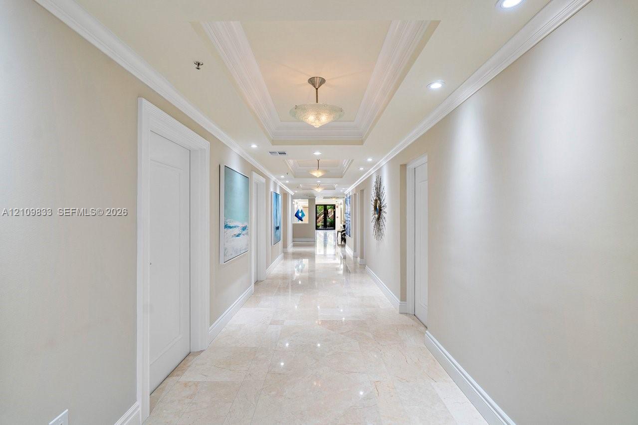Grand gallery hallway featuring stone tile flooring, multi-tiered tray ceilings, recessed lighting, and decorative semi-flush mount fixtures
