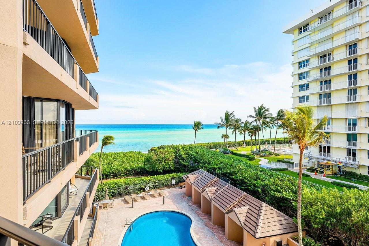 Private balcony with black metal railings overlooking a community swimming pool, poolside cabanas, landscaped grounds, and the ocean Private balcony with black metal railings overlooking a community swimming pool, poolside cabanas, landscaped grounds, and the ocean