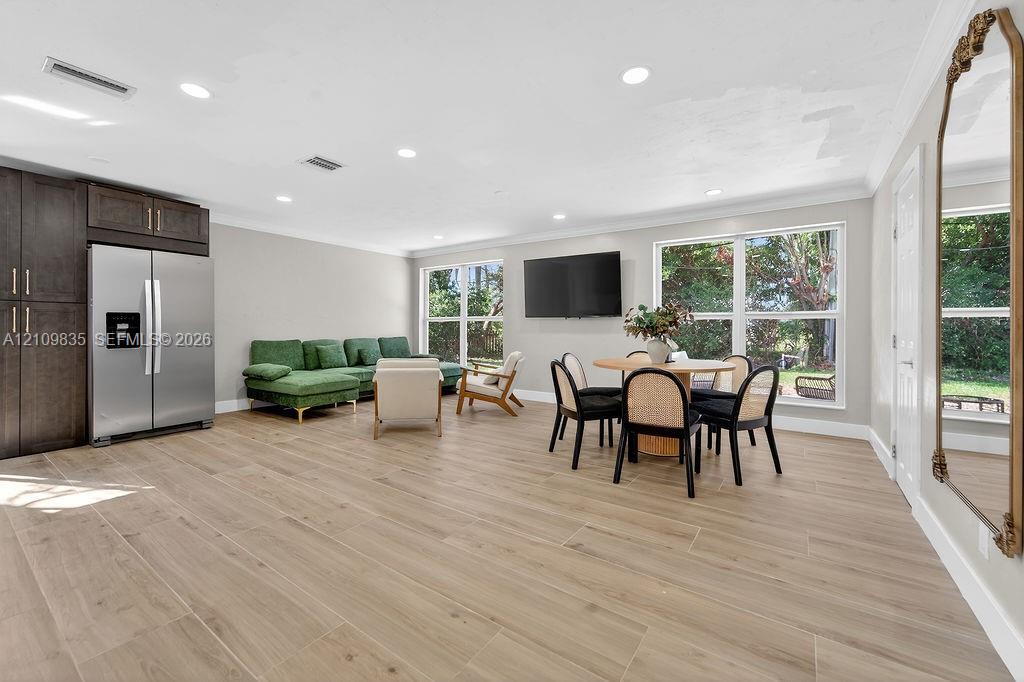 Open-concept space featuring wood-finish flooring, recessed lighting, crown molding, large windows, and built-in dark wood cabinetry with a stainless steel refrigerator