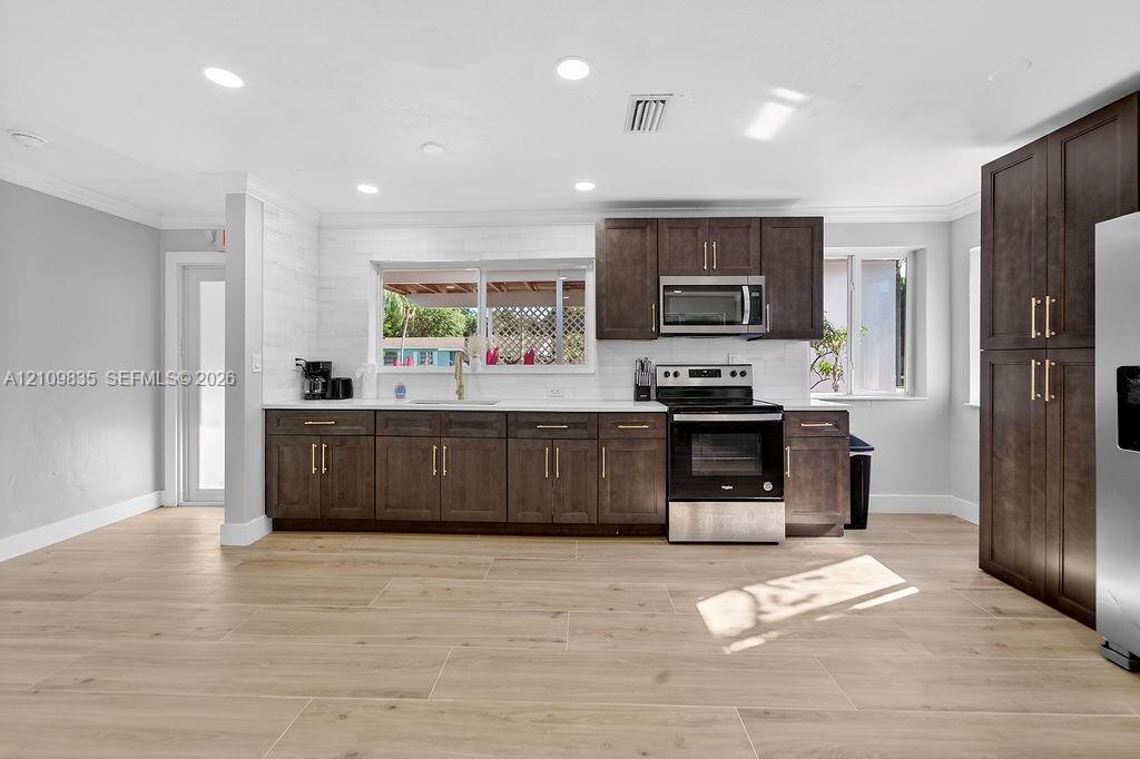 Kitchen featuring dark-toned cabinetry with gold-finish hardware, stainless steel appliances, white subway-style backsplash, light-toned wood-finish flooring, and recessed ceiling lighting