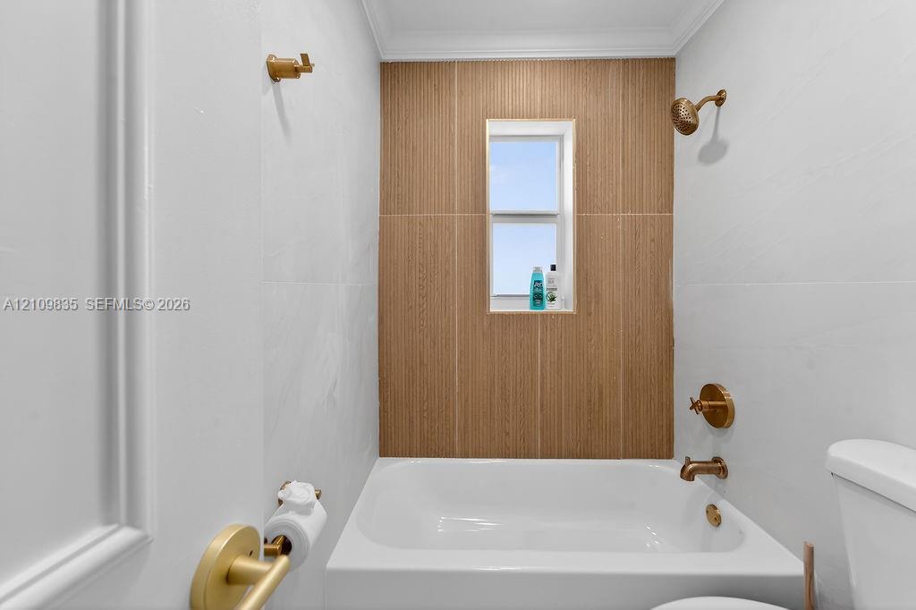 Bathroom featuring white wall tile, a wood-finish tile shower surround, brass-tone fixtures, a built-in tub, and an inset window