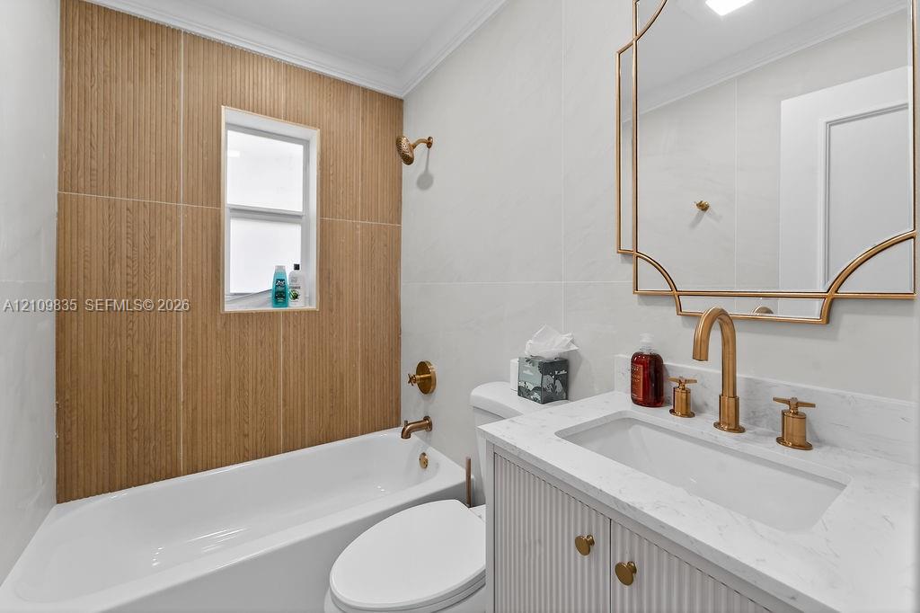 Bathroom featuring a wood-finish tile tub surround, white vanity with stone-style countertop, metallic gold-tone hardware, and decorative wall-mounted mirror