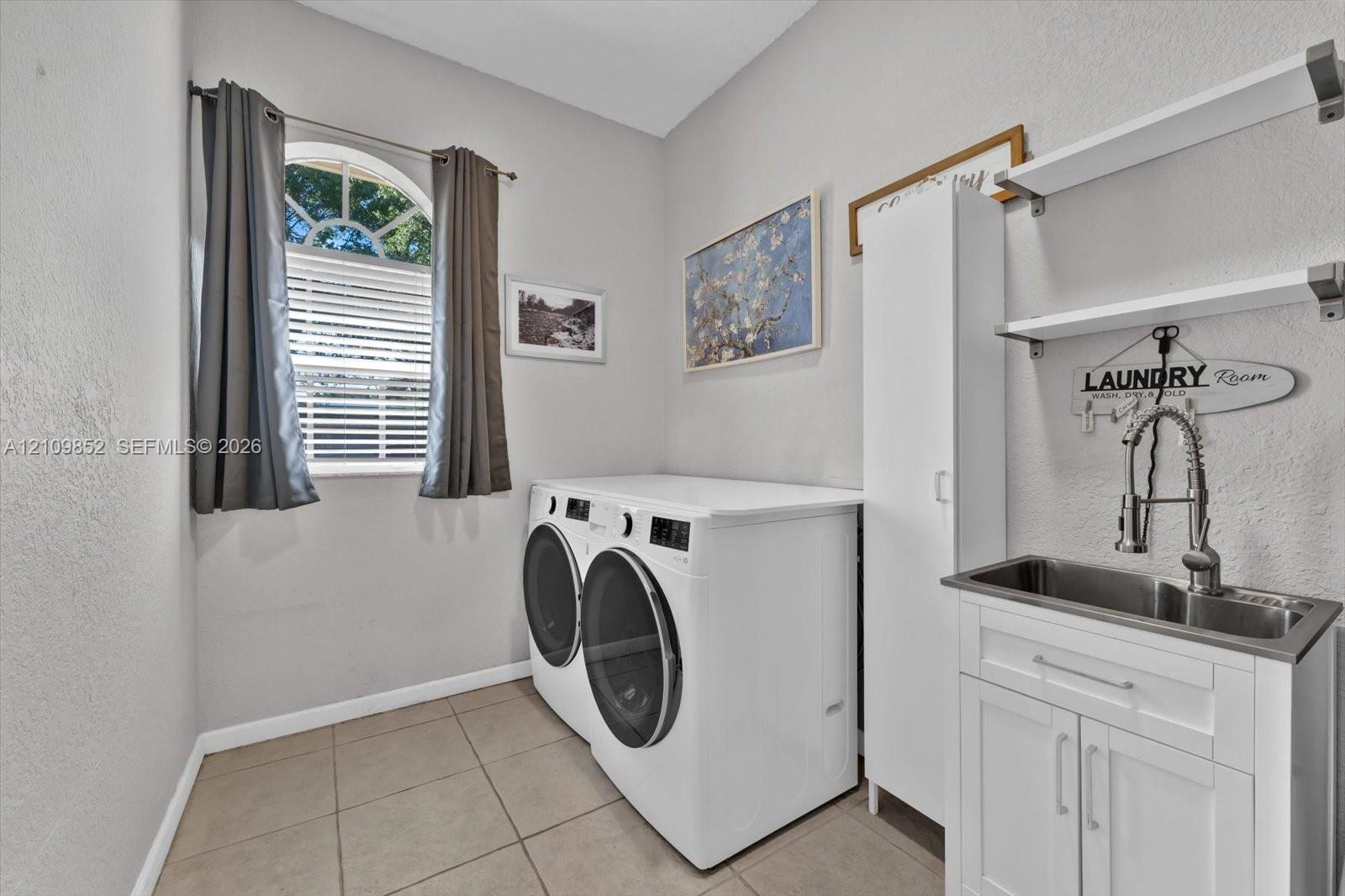 Laundry room featuring ceramic tile flooring, an arched window, a white utility cabinet, and a stainless steel sink with a high-arch pull-down faucet and wall-mounted shelving