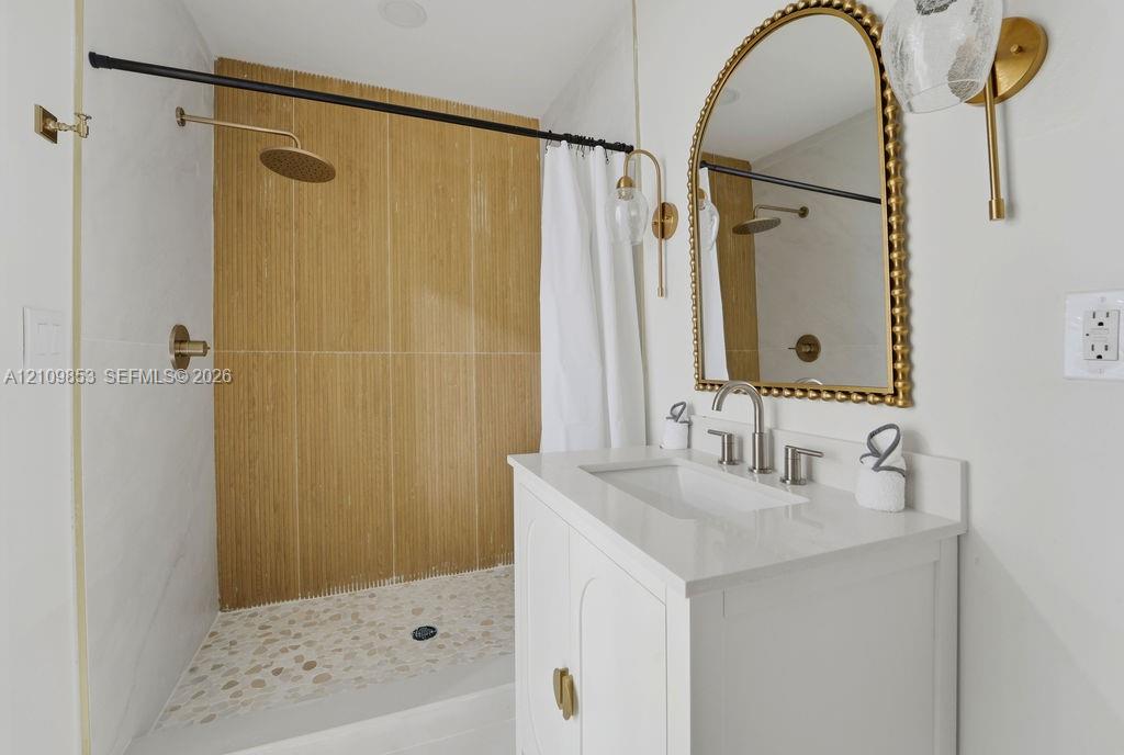 Bathroom featuring a white vanity with a quartz countertop, arched beaded mirror, wall-mounted brass light fixtures, and a shower with wood-tone tile and river stone flooring
