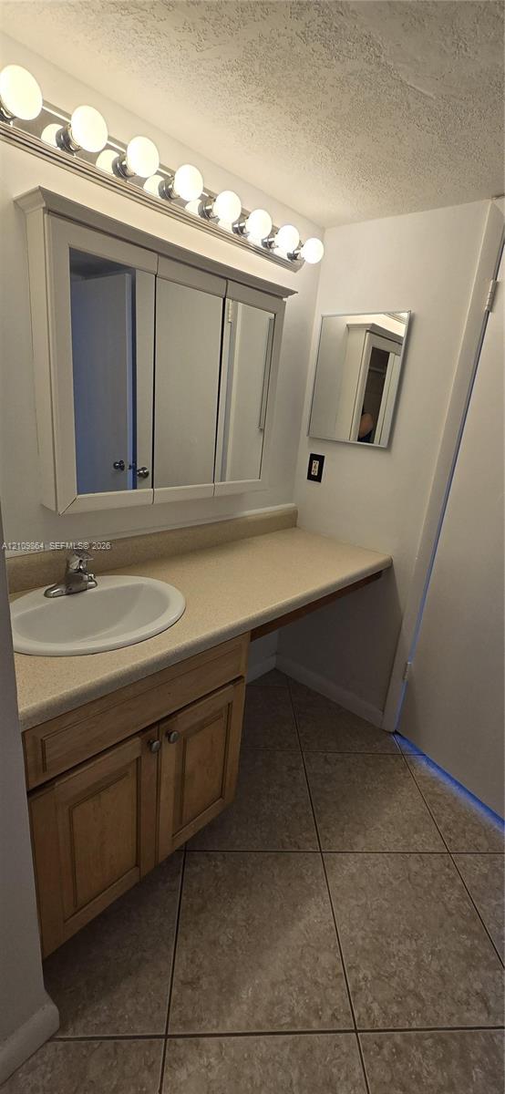 Bathroom vanity featuring light-finish wood cabinetry, a solid-surface countertop with an integrated basin, a tri-fold medicine cabinet, and a multi-bulb light fixture above large-format floor tile
