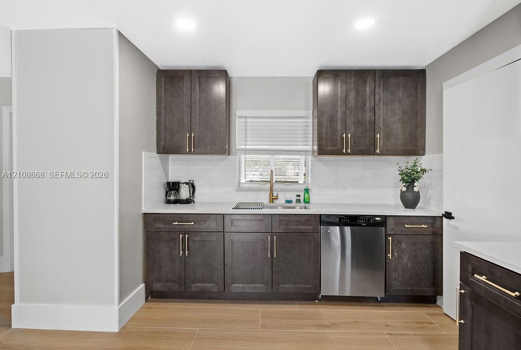 Kitchen featuring dark-finish cabinetry, gold-tone hardware, white stone-surface countertops, subway tile backsplash, and stainless steel dishwasher