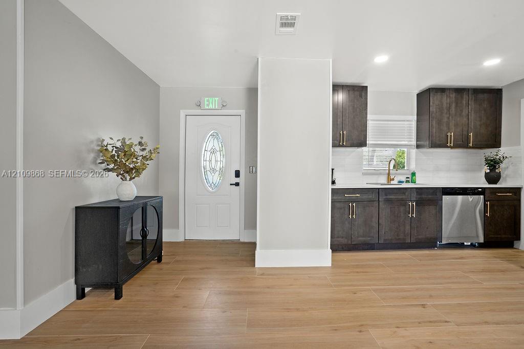 Wood-finish flooring, dark cabinetry with gold-tone hardware, stainless steel dishwasher, recessed lighting, and white door with oval decorative glass insert