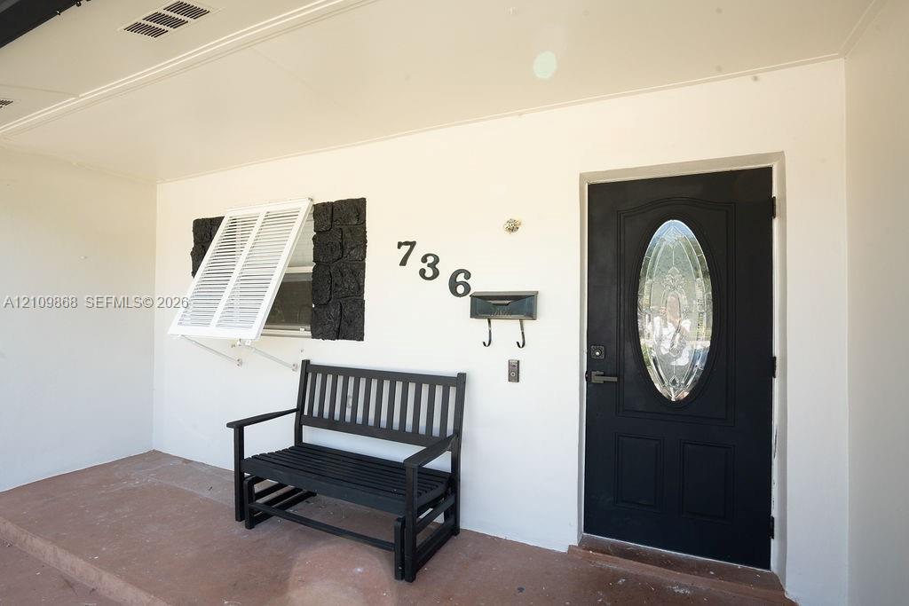 Covered entryway featuring a black front door with an oval glass insert, stone accent wall details, white exterior shutters, and a concrete porch