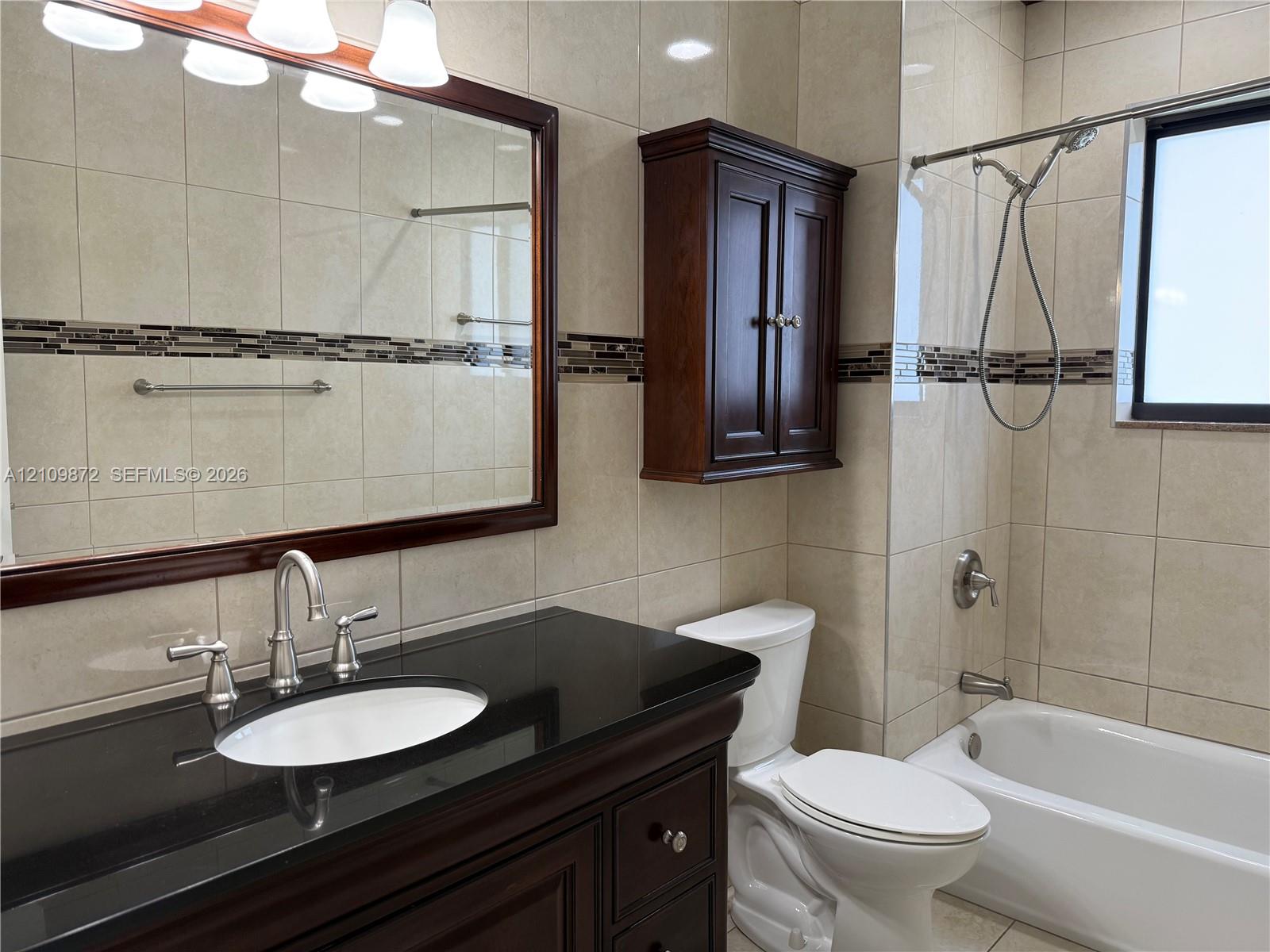 Bathroom featuring a dark wood vanity with a black stone countertop, inset sink, full-width mirror with wood frame, coordinating wall-mounted storage cabinet, and floor-to-ceiling tile with decorative inlay