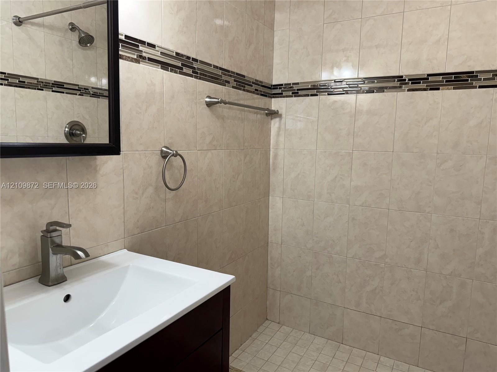 Bathroom featuring a dark wood vanity with a white integrated sink, polished metal faucet, walk-in tiled shower with a decorative mosaic tile inlay, and brushed nickel hardware