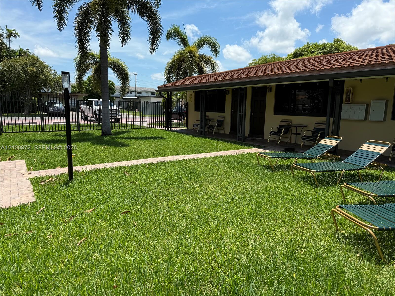 Grassy outdoor space featuring a red-tiled roof, stone-paver walkway, metal perimeter fencing, and mature palm trees
