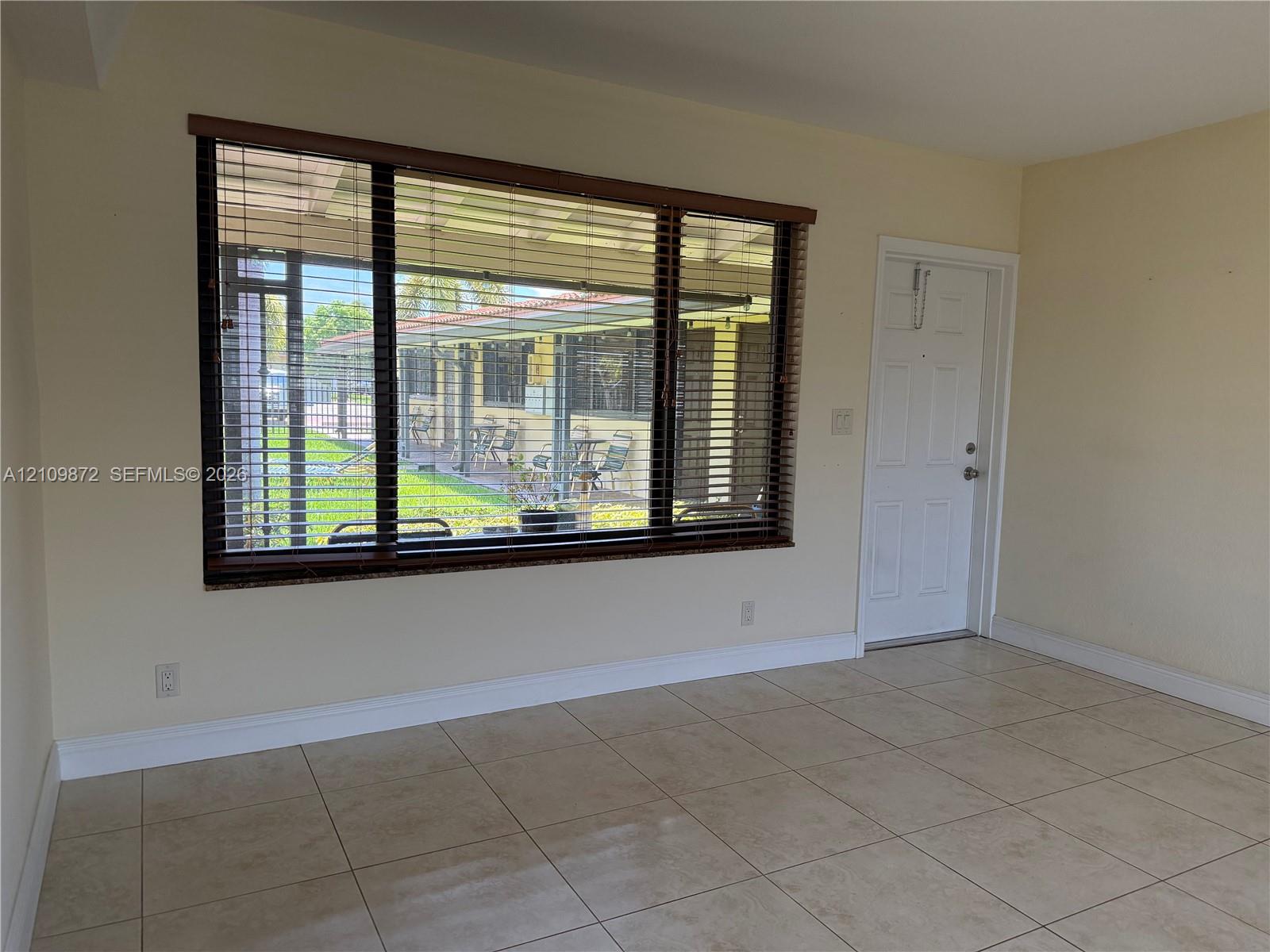 Neutral-toned interior space featuring large-format ceramic tile flooring, a expansive picture window with wood-finish horizontal blinds, and a paneled entry door