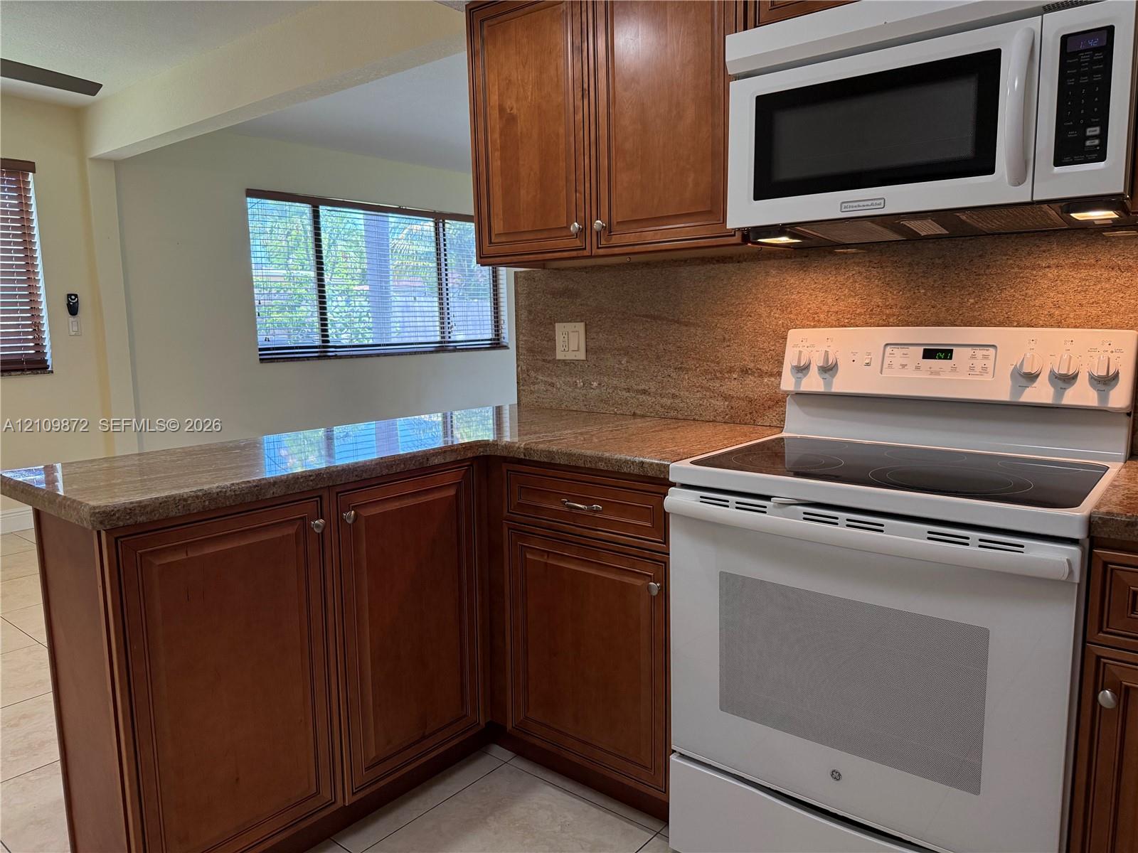 Kitchen featuring stained wood cabinetry, stone-surface countertops, stone-tile backsplash, and white appliances including an electric range and built-in microwave