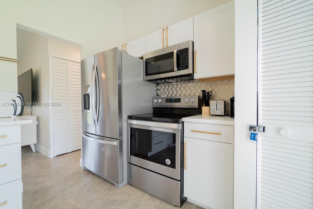 Kitchen featuring white shaker-style cabinetry, stainless steel appliances, brushed gold-tone hardware, a tiled backsplash, and stone-finish countertops