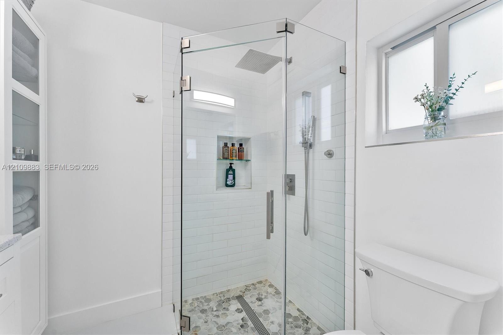 Walk-in glass shower with subway tile surround, integrated shelving, rainfall shower head, and stone-finish mosaic flooring