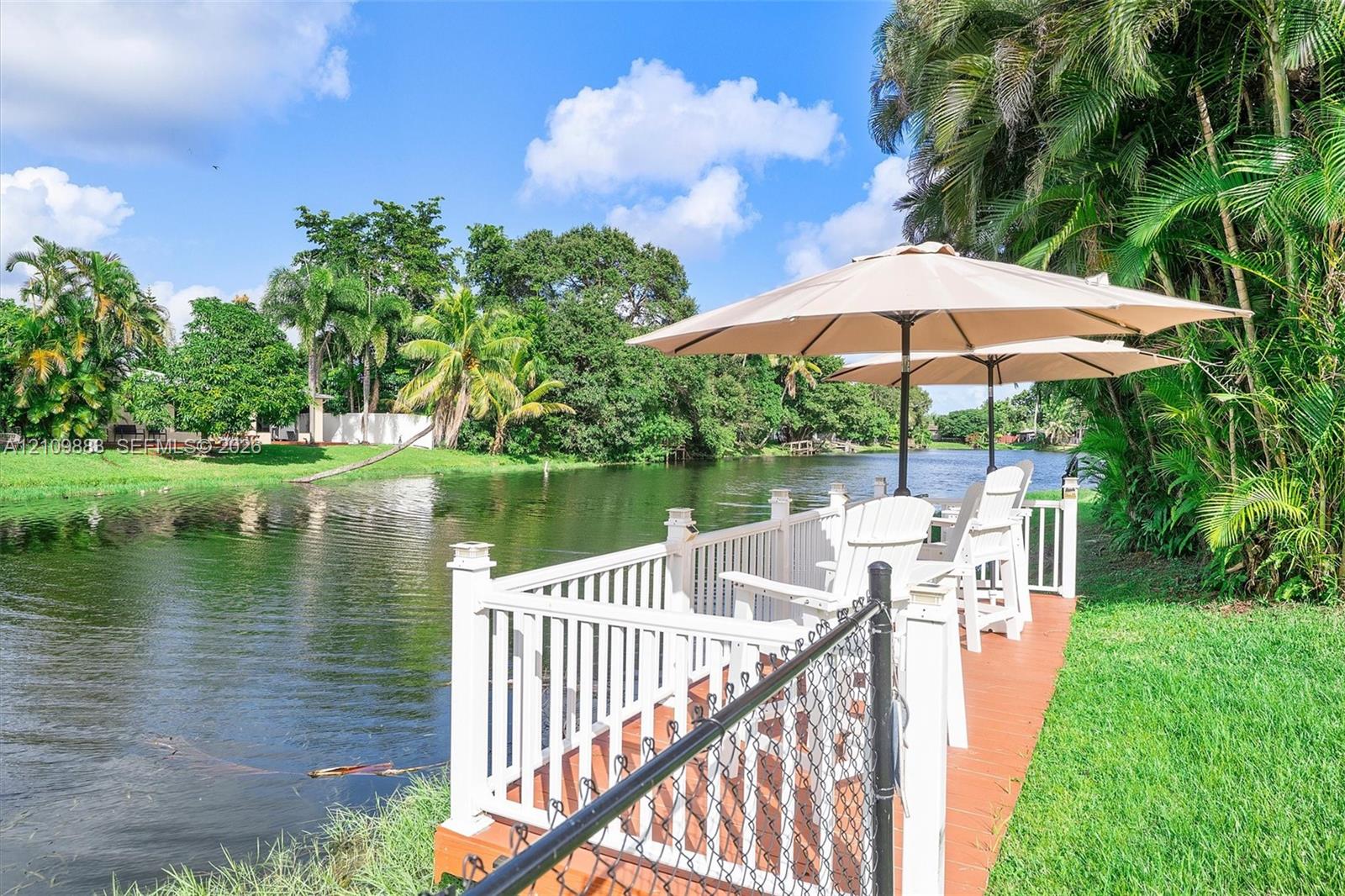 Waterfront deck featuring a wood-tone surface, white railing, and perimeter chain-link fencing, situated along a natural waterway with surrounding tropical landscaping