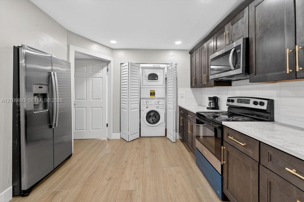 Kitchen featuring dark-stained shaker cabinetry, stainless steel appliances, light-toned stone-finish countertops, wood-finish flooring, and an integrated laundry closet with bi-fold doors